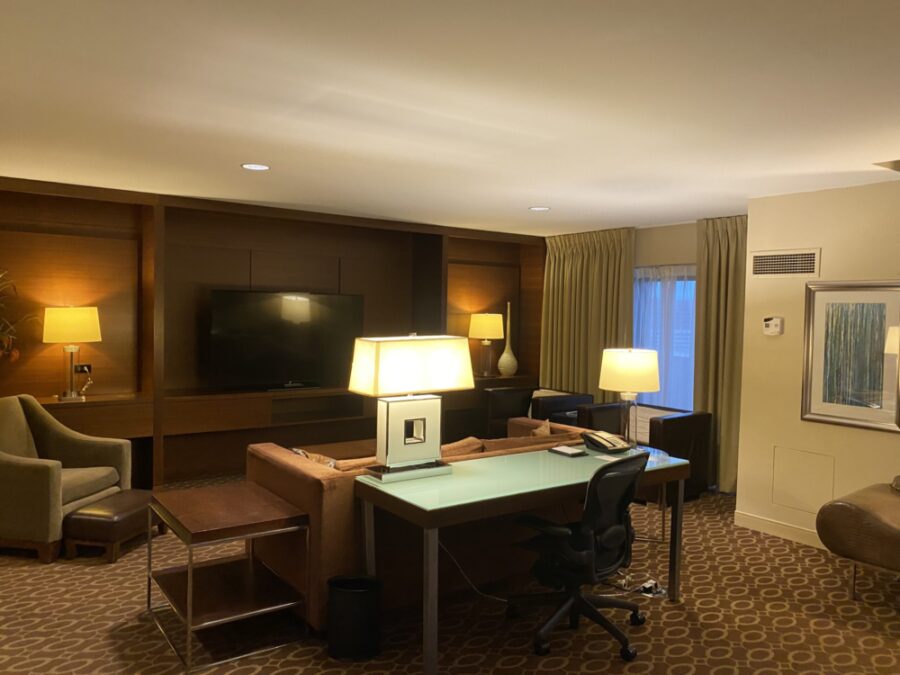 hotel suite with desk behind a coouch, flanked by brown chairs and a tv on the wall. 